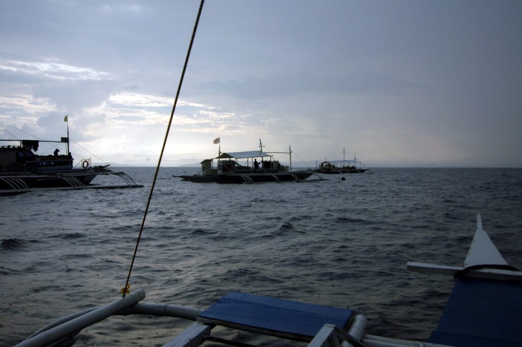 Tři potápěčské lodě banca na kotvě na Monad Shoal pod šedou ranní oblohou, viděné z paluby jiné lodi