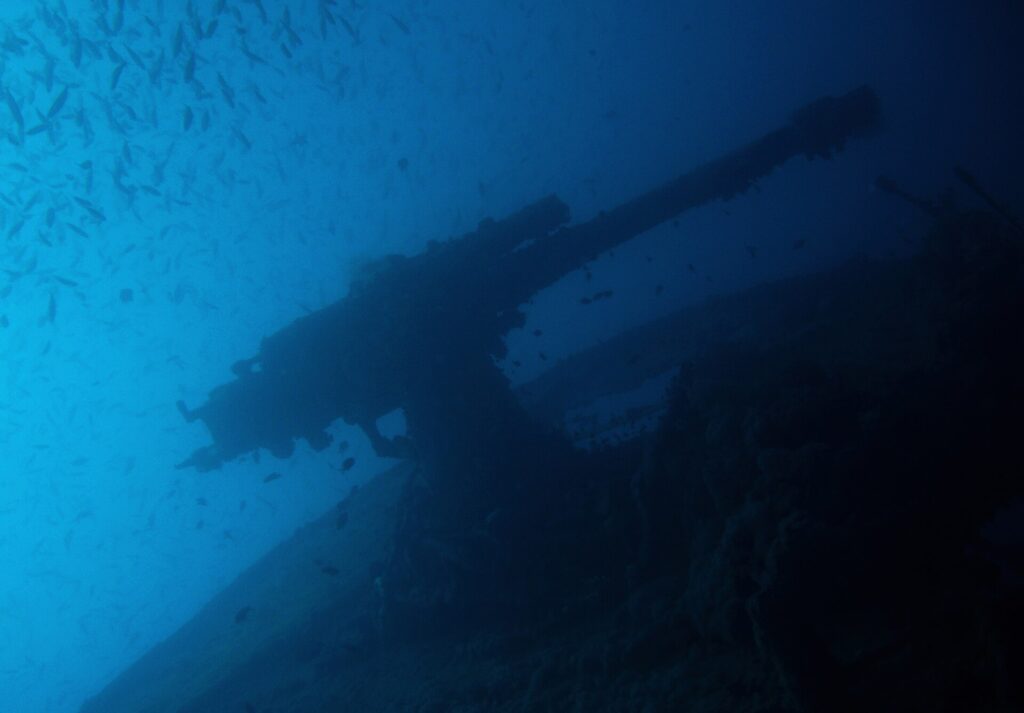 Záďové dělo vraku Thistlegorm jako silueta proti modré, pod hejnem drobných ryb