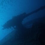 Záďové dělo vraku Thistlegorm jako silueta proti modré, pod hejnem drobných ryb