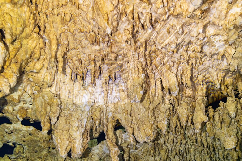 Detail bledě žlutých krápníků a tenkých stalaktitů, dole zvlněný odraz vody