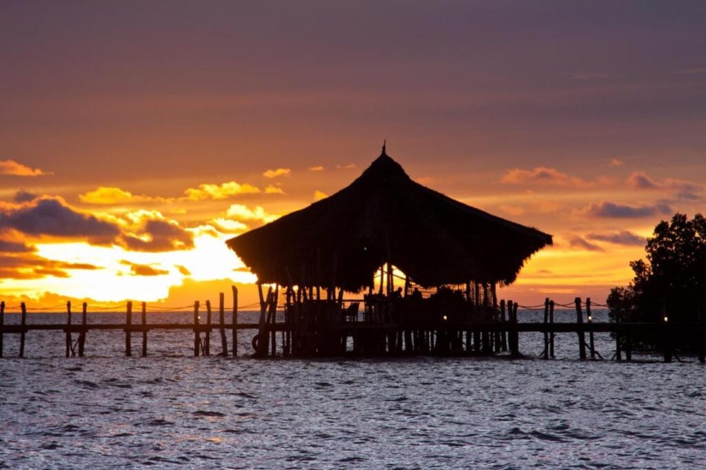 Pavilon se slaměnou střechou na kůlech na konci mola lodge Fundu Lagoon při západu slunce