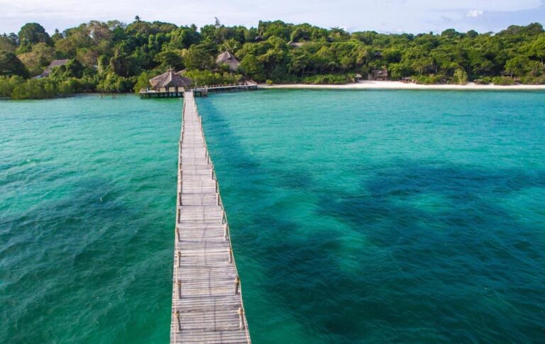 Dlouhé dřevěné molo lodge Fundu Lagoon nad tyrkysovou vodou, naproti zalesněnému pobřeží