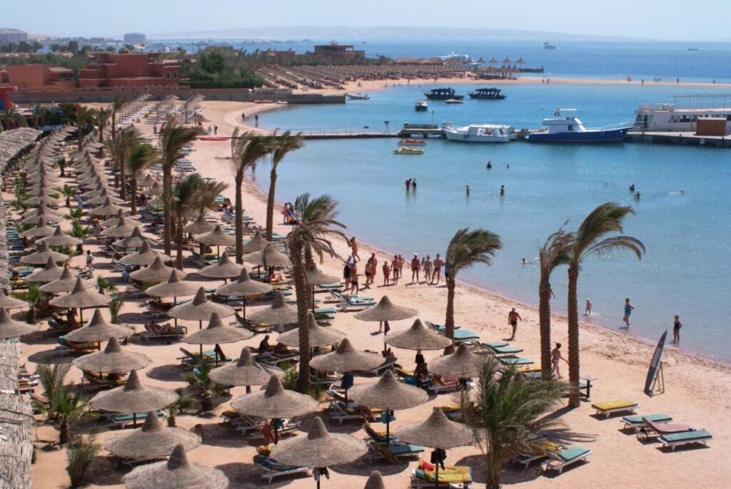Pláž hotelu Giftun Azur Resort se slaměnými slunečníky a palmami, v pozadí molo a lodě