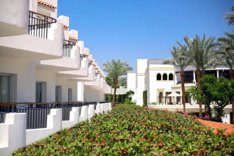 Bílé budovy hotelu JAZ Fanara Resort s balkony a palmami za kvetoucím živým plotem