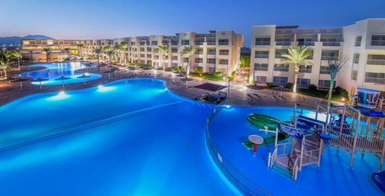 Osvětlené bazény hotelu JAZ Soma Beach za soumraku, mezi budovami a palmami