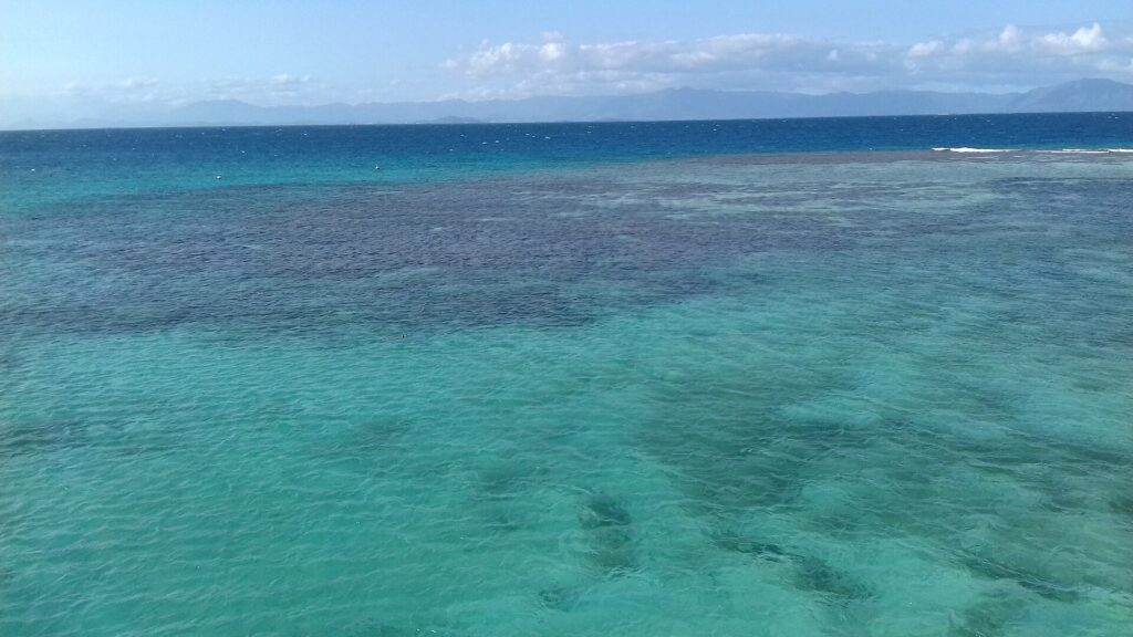 Laguna shora, tyrkysová voda s tmavými skvrnami korálových bloků, vpravo bariéra, o kterou se lámou vlny, a na obzoru hory ostrova Grande Terre