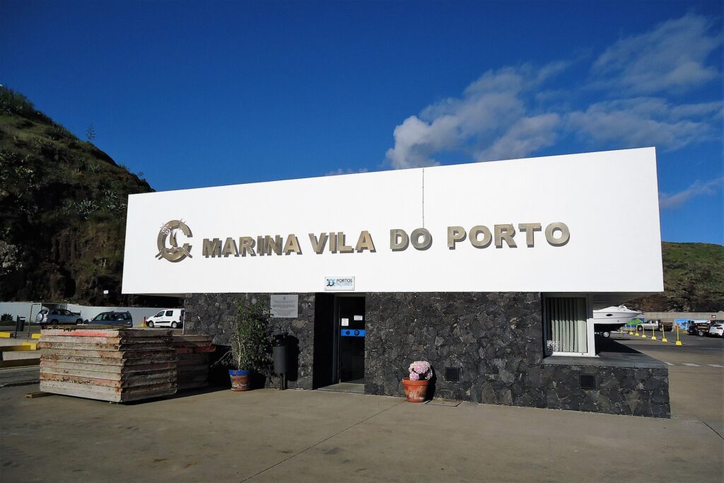 Recepce mariny ve Vila do Porto, velký bílý nápis z kovových písmen na zdi z černého čediče, pod modrou oblohou, vlevo zelený sráz