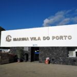 Recepce mariny ve Vila do Porto, velký bílý nápis z kovových písmen na zdi z černého čediče, pod modrou oblohou, vlevo zelený sráz