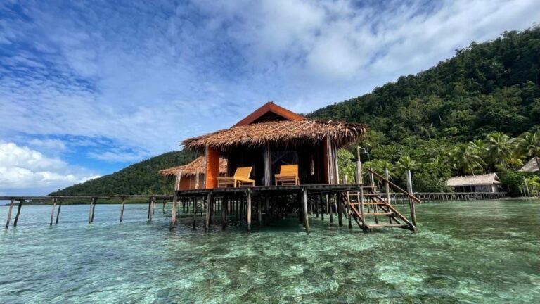 Bungalov penzionu Nyande Raja Ampat na kůlech nad průzračnou vodou, pod zalesněným kopcem