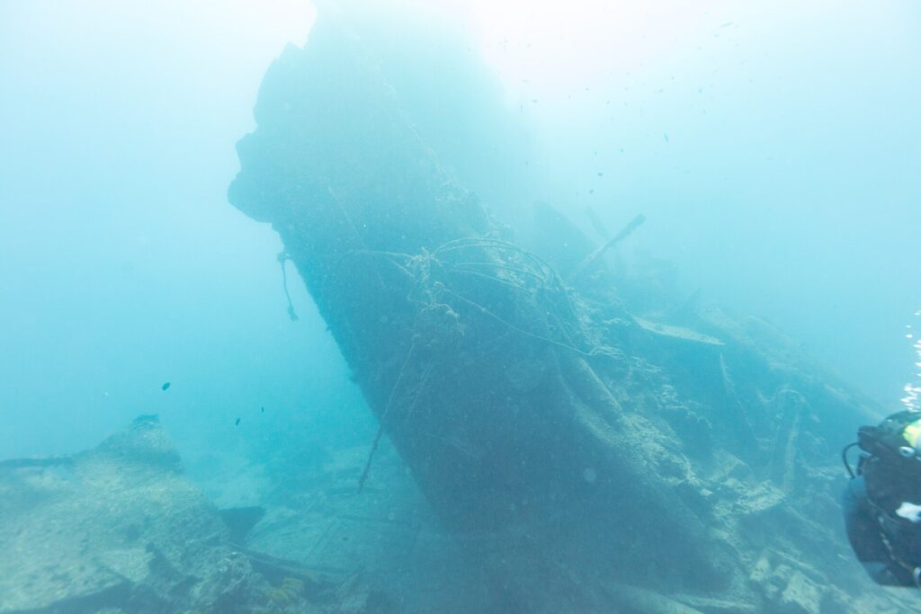 Zlomená záď vraku Thistlegorm se zvedá nakloněná do modré, vpravo potápěč