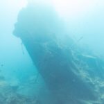 Zlomená záď vraku Thistlegorm se zvedá nakloněná do modré, vpravo potápěč