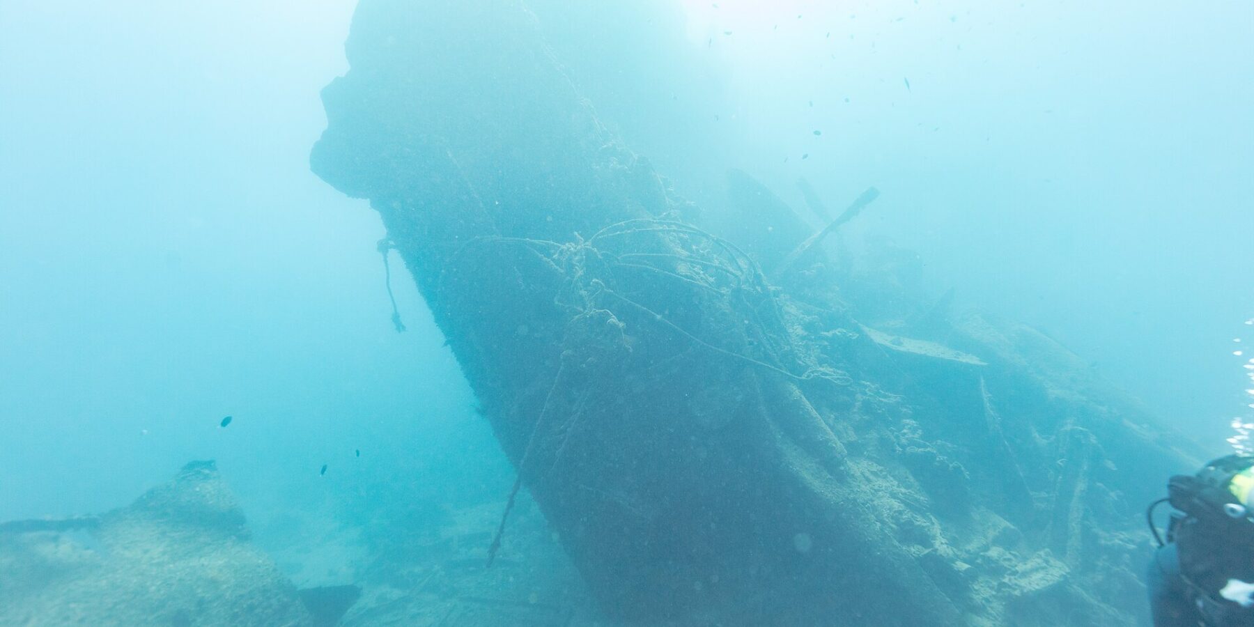 Zlomená záď vraku Thistlegorm se zvedá nakloněná do modré, vpravo potápěč