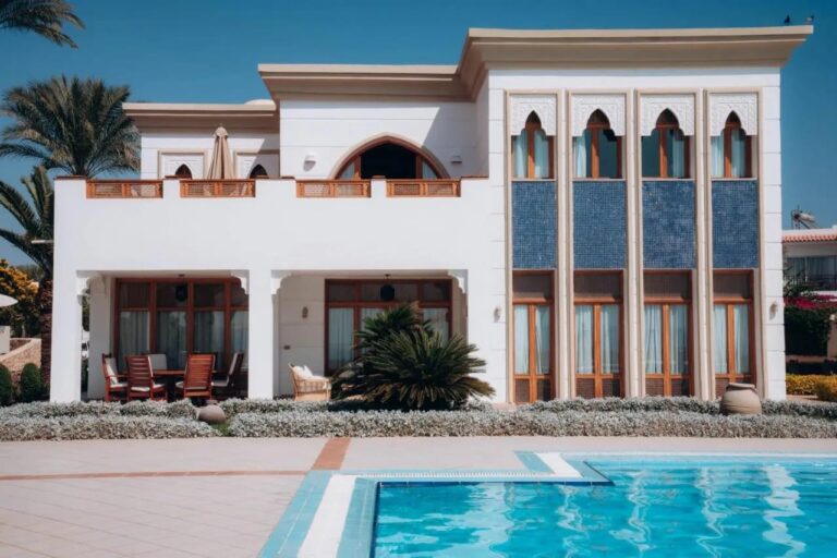 Bílá budova Reef Oasis Beach Resort s obloukovými okny a panely z modré mozaiky, před bazénem