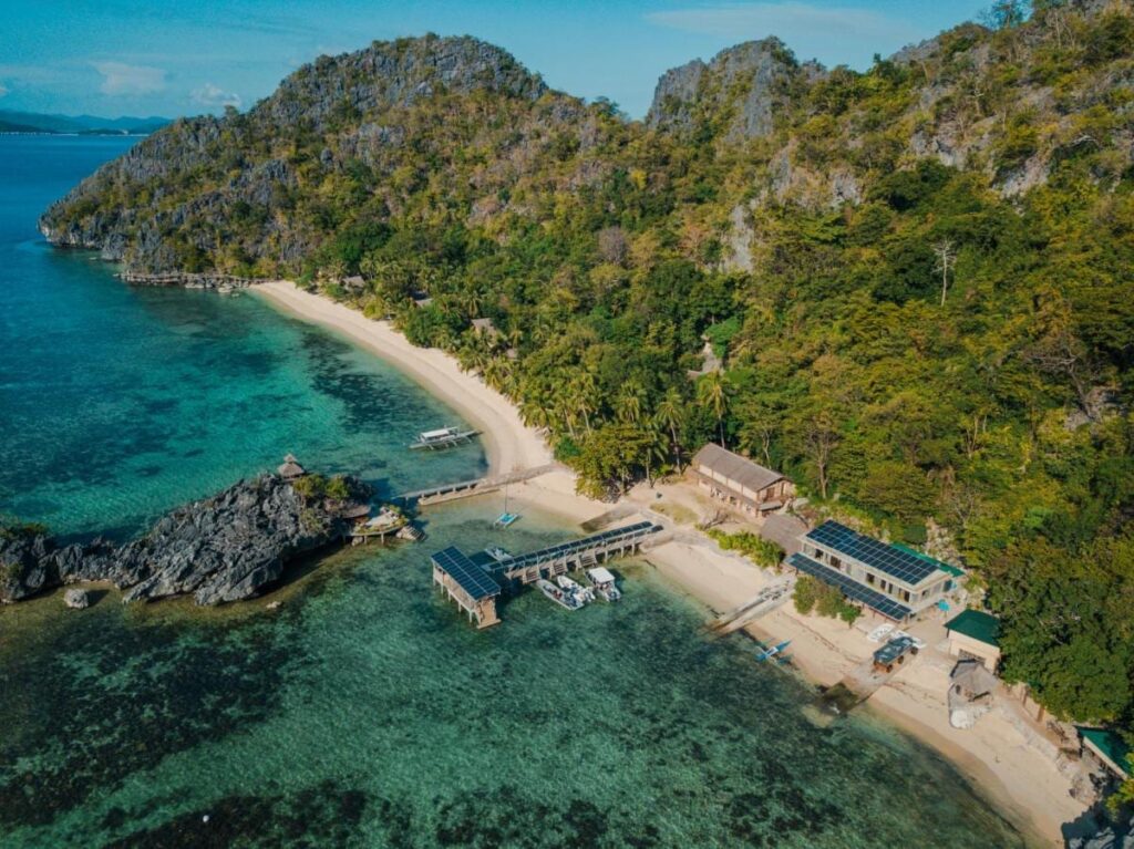 Letecký pohled na Sangat Island Dive Resort, jeho pláž, molo a lodě pod skalními stěnami