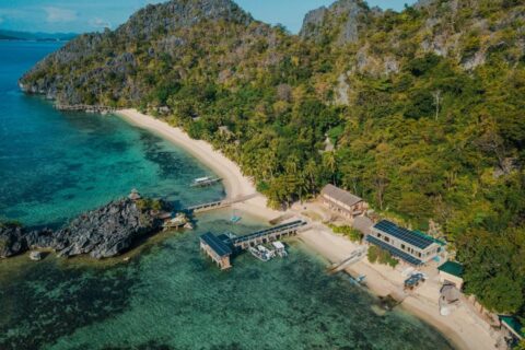 Letecký pohled na Sangat Island Dive Resort, jeho pláž, molo a lodě pod skalními stěnami