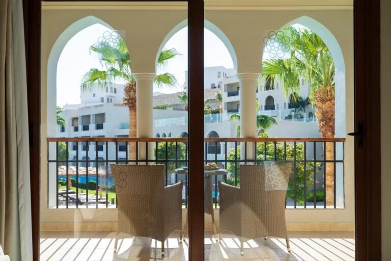 Balkon s arkádami a dvěma pletenými křesly, s výhledem na palmy a bílé budovy Sunrise Arabian Beach Resort