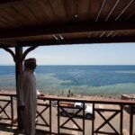 Muž v galabíji se z dřevěné terasy dívá na jámu Blue Hole lemovanou světlým útesem, kde plavou koupající se lidé