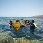 Plavci ve žlutých vestách, s maskami a šnorchly v čiré vodě Blue Hole, za nimi hory Sinaje